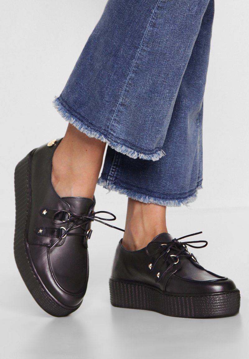 Chaussures à plateforme noires à lacets portées avec un jean bleu court à ourlet effiloché, pieds croisés.