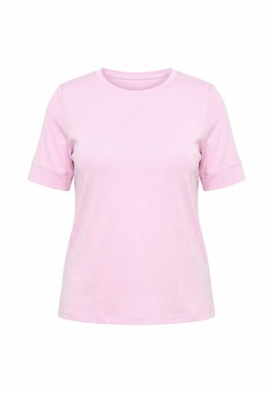 Femme aux cheveux blonds ondulés, portant un t-shirt rose clair à manches courtes et un pantalon vert, debout avec une main sur la hanche, de dos.