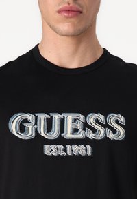 Juoda medvilninė marškinėliai su dideliu, tekstūruotu siuvinėjimu, kuriame parašyta "GUESS" mėlyna ir balta spalvomis, o po juo nurodyta "EST. 1981".