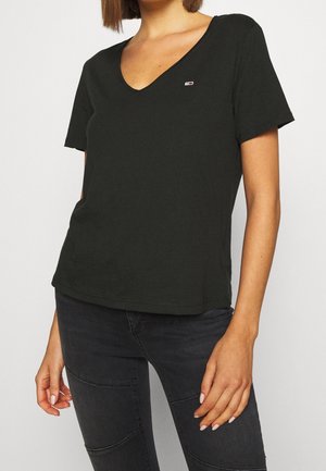 Frau, die ein schwarzes V-Ausschnitt T-Shirt mit kurzen Ärmeln und eng anliegende schwarze Jeans trägt, mit sichtbarem Oberkörper und oberen Beinen vor einfarbigem Hintergrund.