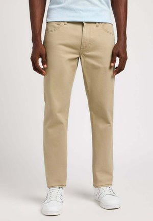 Pantalones de algodón beige con un diseño de pierna recta. Cuenta con el clásico estilo de cinco bolsillos y un cierre de cremallera. Combinado con zapatillas blancas.