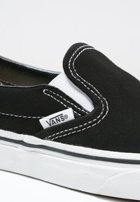 Baskets Vans slip-on noires avec couture blanche, accents élastiques sur les côtés et semelle en caoutchouc blanche avec détail de bande noire.