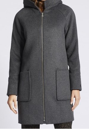 Kvinde iført en knælængde mørkegrå hættetrøje med lynlås foran og to store rektangulære påsyede lommer.