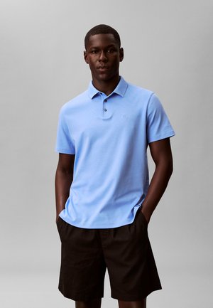 Man draagt lichtblauw poloshirt met korte mouwen en zwarte korte broek, staat met handen in de zakken tegen een neutrale grijze achtergrond.