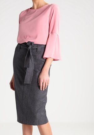 Femme portant un chemisier rose à manches évasées et une jupe crayon gris foncé taille haute avec des boutons à l'avant et une ceinture en tissu nouée.