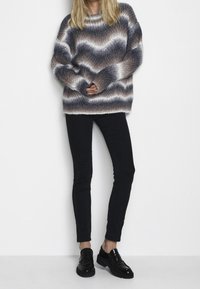 Oversized gebreide trui met een golvend patroon in grijs, bruin en wit, gecombineerd met zwart slankzittende broek en zwarte chunky loafers.