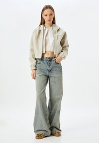 Beiger, taillierter Hoodie mit Kordelzügen, über einem weißen Cropped-Top getragen. Kombiniert mit hellblauen, weit geschnittenen Jeans und Schuhen mit Leopardenmuster.
