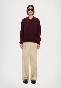 Burgunderfarbener Strickpullover mit Kragen und drei Knöpfen, kombiniert mit cremfärbigen, gestreiften weit geschnittenen Hosen und schwarzen klobigen Schuhen.