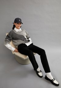 Grijze gestreepte trui over een wit shirt, zwarte broek en zwarte schoenen met witte accenten. Zittend op een luxe beige stoel tegen een grijze achtergrond.