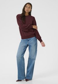 Pull en tricot bordeaux avec des manches plissées, associé à un jean ample bleu clair et des escarpins bordeaux à bout pointu. Fond lumineux.