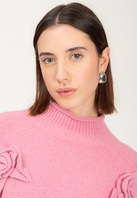 Giovane donna con capelli castani fino alle spalle, che indossa orecchini d'argento e un maglione rosa a trama con dettagli a forma di rosa sulle spalle.