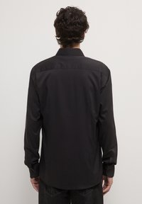 Maglietta nera a maniche lunghe con una texture liscia, polsini con bottoni e colletto classico. La vista posteriore mostra un design semplice e su misura.