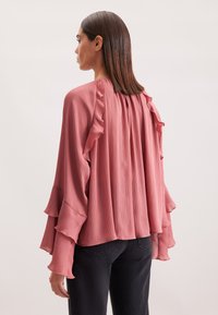 Blusa plissettata di un rosa tenue con maniche arricciate e dettagli sul retro, in un tessuto leggero, indossata con jeans scuri, che mette in risalto la texture e il movimento.