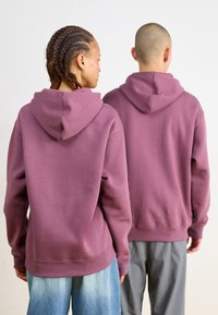 Dve postavy nosia zladene oversized mikiny v jemne fialovej farbe. Tkanina vyzerá mäkká, s klokankou a rebrovanými manžetami.