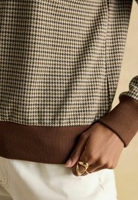 Brown and cream houndstooth patterned top with ribbed brown cuffs, featuring a relaxed fit and textured fabric. Hand resting on white pants.