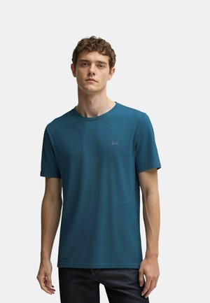 Giovane uomo con capelli corti e ricci, indossa una maglietta a maniche corte teal e pantaloni scuri, in piedi contro uno sfondo chiaro uniforme.