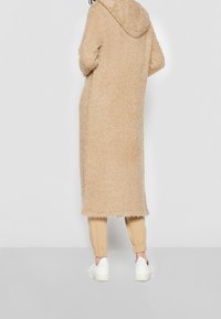 Beige, pörröinen pitkä takki hupulla, jossa kuvioitu kangas. Päällä vaaleanbeigejen verryttelyhousujen ja valkoisten lenkkareiden kanssa, joissa on joustavat kalvonauhat.