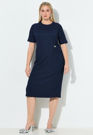 Donna bionda che indossa un vestito blu navy al ginocchio con maniche corte e décolleté argento con fiocchi, in piedi contro uno sfondo bianco.