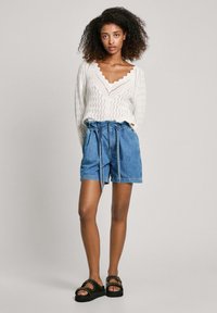 Suéter blanco con escote de ondas, combinado con pantalones cortos de mezclilla azul que tienen una cinturilla elástica y cordones. Sandalias negras con tachuelas.