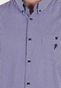 Chemise à carreaux bleus et blancs avec un col boutonné, boutons noirs et une poche poitrine arborant un petit logo. Matière en mélange de coton.