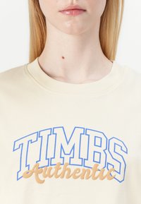 Kremasta pamučna majica s okruglim izrezom i plavim i smeđim natpisom "TIMBS Authentic" otisnutim na prednjoj strani.