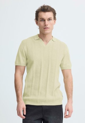 Man met lichtbeige gebreide polo met korte mouwen en zwarte broek, staand tegen een lichte, effen achtergrond, kijkend naar voren.