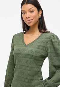 Pull en tricot vert texturé avec un col en V, des manches ballon et de subtiles rayures horizontales. Fabriqué en tissu léger avec un design en maillage ouvert.