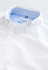 White button-down shirt with blue striped inner collar and "Next Regular" size label, laid flat on a white surface.