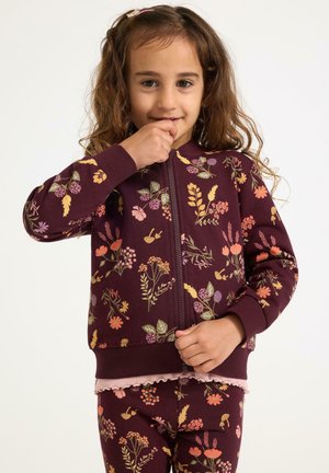 Ragazzina con lunghi capelli ricci che indossa una giacca zip-up floreale bordeaux e pantaloni abbinati, tenendo la mano vicino alla bocca.