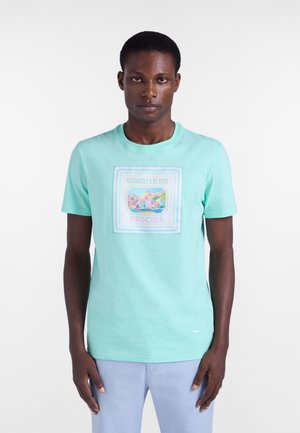 Homme portant un t-shirt graphique bleu clair avec le texte « Harmont & Blaine Procida » et un design pastel de paysage urbain, debout sur un fond blanc.