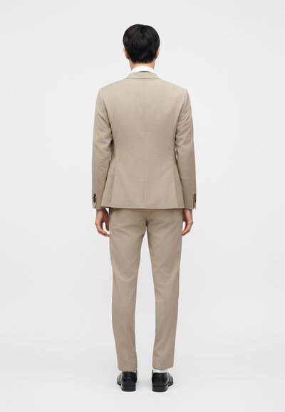 Μπεζ κοστούμι που περιλαμβάνει ένα ραμμένο σακάκι και τζιν παντελόνια που ταιριάζουν, με λεία υφή και κλασική κοπή. Μαύρα παπούτσια ολοκληρώνουν την εμφάνιση.