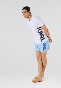 T-shirt blanche avec impression noire "KARL" sur le côté, associée à un short de bain bleu clair arborant un logo "KARL" bleu. Tissus en coton et nylon lisses.