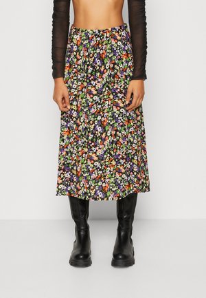 Kobieta w midi spódnicy w kwiaty w kolorach fioletowym, pomarańczowym i białym, w połączeniu z czarnymi kozakami za kolano i przezroczystą bluzką z długim rękawem.