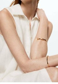 Bracelets en or sur des bras nus, avec une finition lisse et brillante et un design minimaliste. Blouse blanche légère et texturée avec un col.