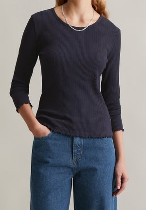 Femme portant un haut côtelé bleu marine à manches longues avec un ourlet ondulé et un jean bleu, main dans la poche, devant un fond clair uni.