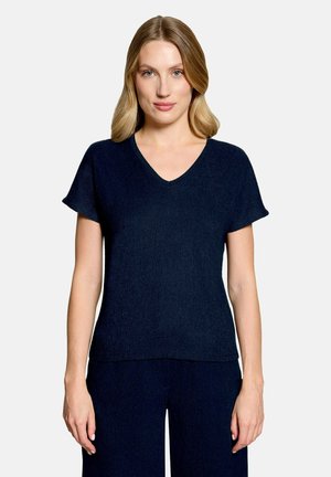 Frau mit blondem Haar, die ein marineblaues V-Ausschnitt-Kurzarmshirt und passende Hosen trägt, steht vor einem schlichten hellen Hintergrund.