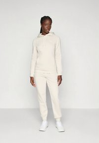 Svetlo bež pulover s kapuco in vrvico, kombiniran z ujemajočimi se ohlapno zasukanimi trenirkami. Ima prefinjeno majhno logotip na sprednji strani in elastične manšete.