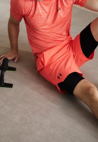 Idrottare i en strukturerad, kortärmad, ljusorange tröja och matchande shorts över svarta kompressionsbyxor, som vilar på en slät yta.