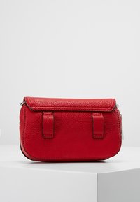 Petite pochette en cuir rouge avec fermeture à rabat et deux sangles cousues à l'avant, présentée sur un socle blanc.