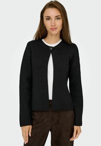 Schwarze Strickjacke mit rundem Ausschnitt, Knopfverschluss am Kragen und langen Ärmeln; strukturiertes Material, über einem weißen Shirt getragen.