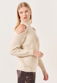 Pull en maille côtelée beige avec un col montant et un design épaules nues. Manches longues avec un tissu texturé et un ourlet ajusté. Motif texturé sur le pantalon.