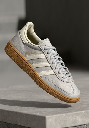 Lichtgrijze suède Adidas Spezial sneaker met witte strepen en gumzool, geplaatst op de rand van een betonnen oppervlak.