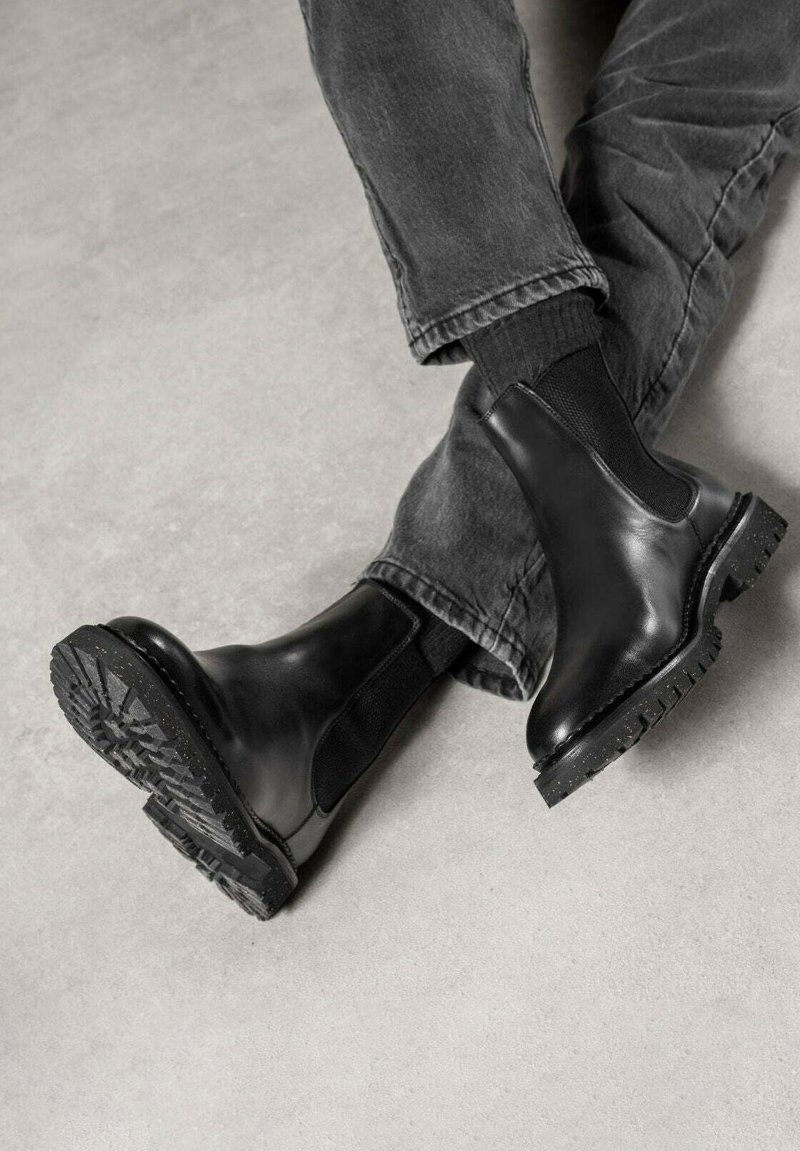 Bottes Chelsea en cuir noir avec panneaux latéraux élastiques et semelle texturée à crampons robustes, portées avec un pantalon gris.