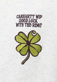 Hellgrauer Stoff mit gesticktem grünem vierblättrigem Kleeblatt und lila Schrift: "CARHARTT WIP GOOD LUCK WITH THE REST."
