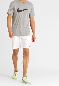 Grå kortärmad Nike-t-shirt med svart logotyp, kombinerat med vita shorts och grå sneakers med neonkanton och svarta detaljer.