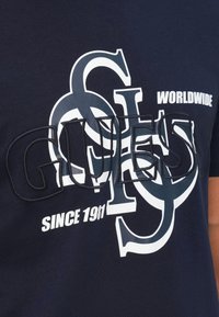 Camiseta de algodón azul marino con un gran logotipo entrelazado de la "G" y la "S" en blanco y azul claro, con acentos de texto que dicen "MUNDIAL" y "DESDE 1981".