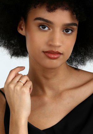 Goldener Ring mit zwei klaren Edelsteinen, getragen an einer Hand mit heller Haut. Das Subjekt hat dunkles, lockiges Haar und trägt ein schwarzes Oberteil.