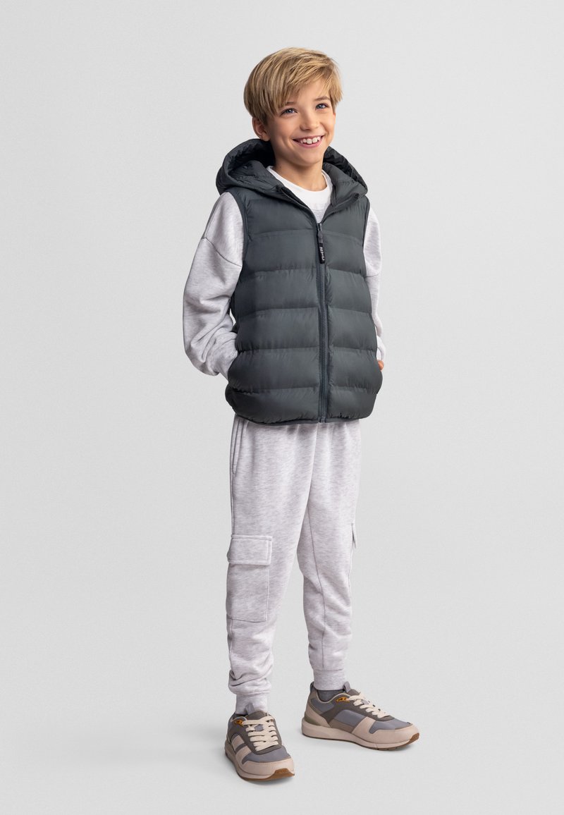 Niño sonriente con las manos en los bolsillos, lleva un chaleco con capucha gris, pantalones deportivos gris claro y zapatillas grises, de pie sobre un fondo liso gris claro.