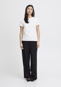 White cotton t-shirt with a small multicolored logo on the left chest, paired with black, loose-fitting, linen pants.