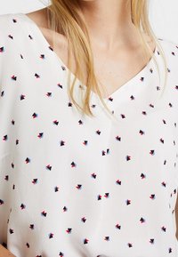 Blusa de textil blanca con escote en V, que presenta un patrón de pequeñas formas geométricas en rojo, azul y negro. Tela suave y ligera.